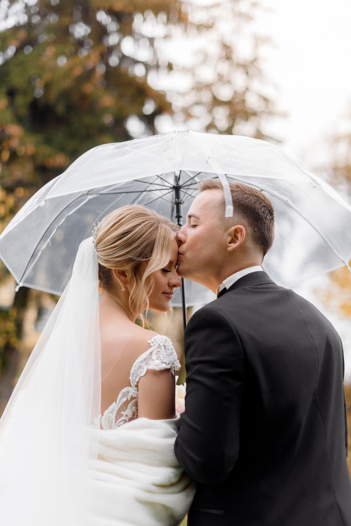 Back view of young groom kisses a blonde bride under an umbrella