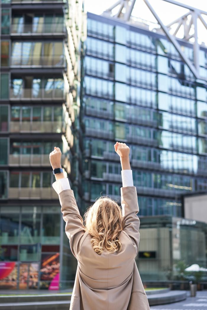 Rear view of corporate woman, lawyer celebrating, lifting hands up and triumphing, achieve goal or success, standing outside on street of city center