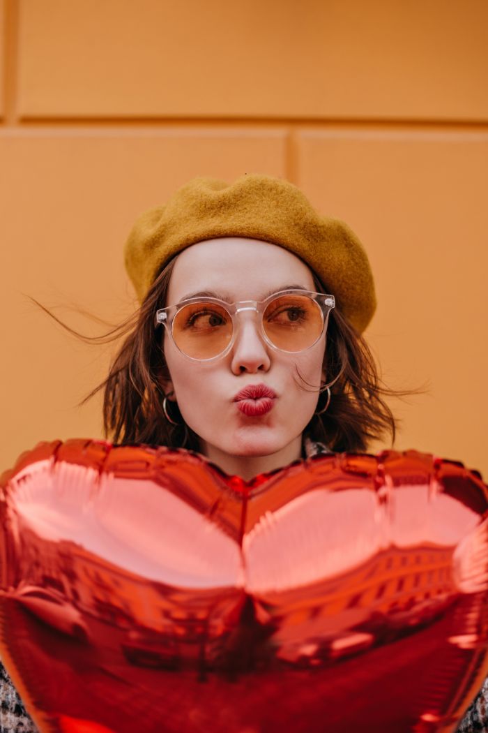 Young girl in yellow beret sends kiss. Woman hiding behind red heart-shaped balloon