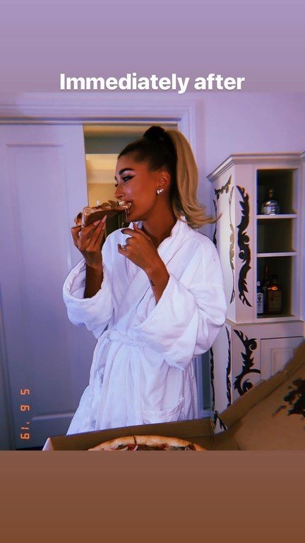 H Hailey Bieber φαίνεται στο δωμάτιο ενός ξενοδοχείου να φοράει το μπουρνούζι της και να τρώει ένα κομμάτι πίτσα.