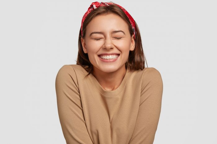 Portrait of happy European woman has broad smile, closes eyes, feels excitement, being in high spirit, recieves proposal from boyfriend, laughs at something funny, models against white background