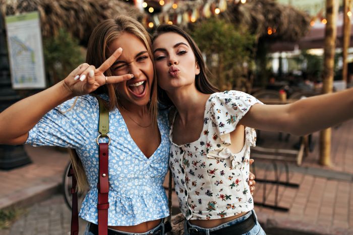 Crazy happy blonde woman in blue floral blouse winks, widely smiles and shows peace sign. Brunette