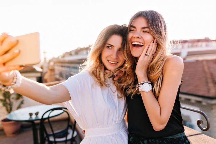 Portrait of laughing friends enjoying weekend together and making selfie on morning city background. Gorgeous woman taking picture of herself standing near smiling friend wearing stylish bracelet..