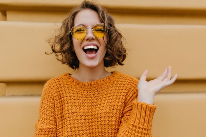 Enthusiastic smiling girl with shiny curls posing in front of old wall. Close-up outdoor portrait of enchanting lady in sweater and trendy glasses..