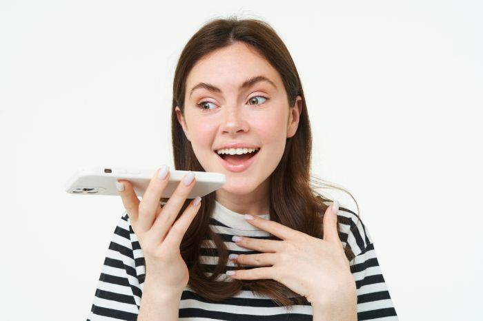 Image of cute brunette woman talking into speakerphone, holding mobile phone near mouth, records her voice, sends a voicemessage, using online translator app, white background