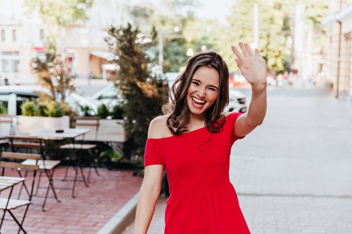 Happy woman in trendy red dress waving hand. Outdoor shot of pos