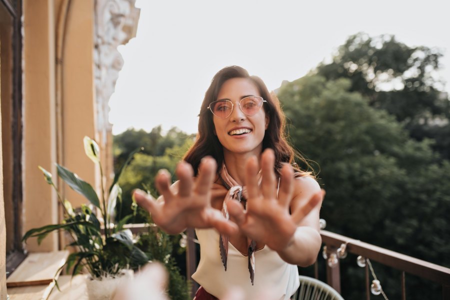 Young girl in sunglasses reaches out to camera. Beautiful woman with dark wavy hair in white blouse is smiling and posing on terrace.