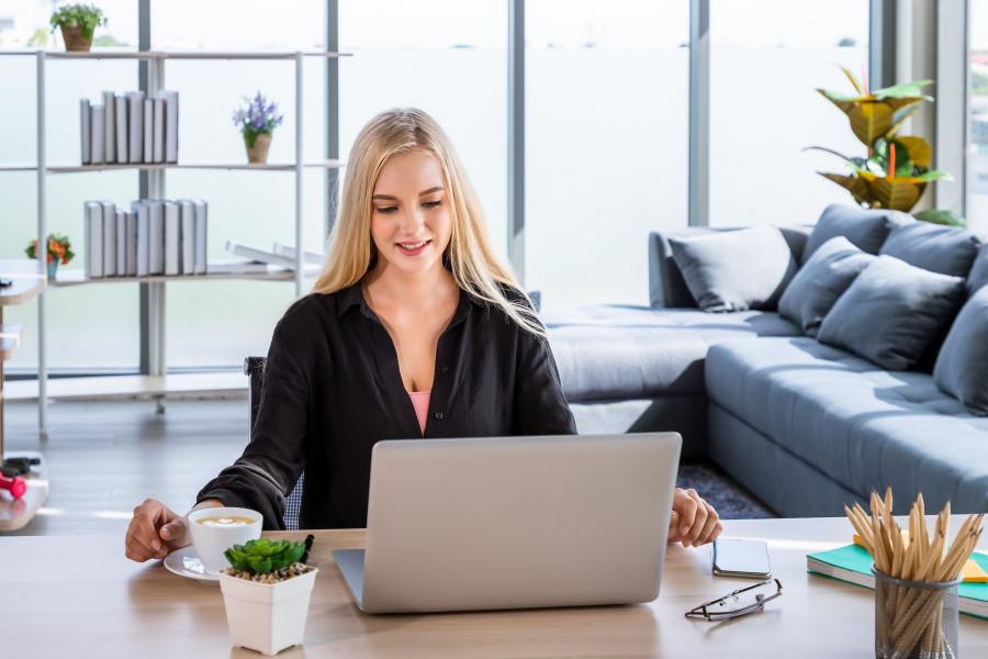 Portrait of young Caucasian woman with blonde hair freelancer working at home, sitting at desk in living room using typing laptop computer