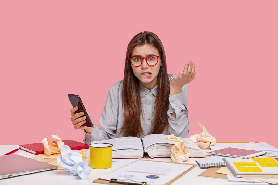Stressful woman bites lower lip, raises hand, types text message on cell phone, tries to find unknown word in online dictionary, poses at workplace with paper crags, drinks hot tea or coffee