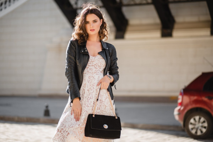 attractive woman walking in street in fashionable outfit, holding purse, looking down, wearing black leather jacket and white lace dress, spring autumn style