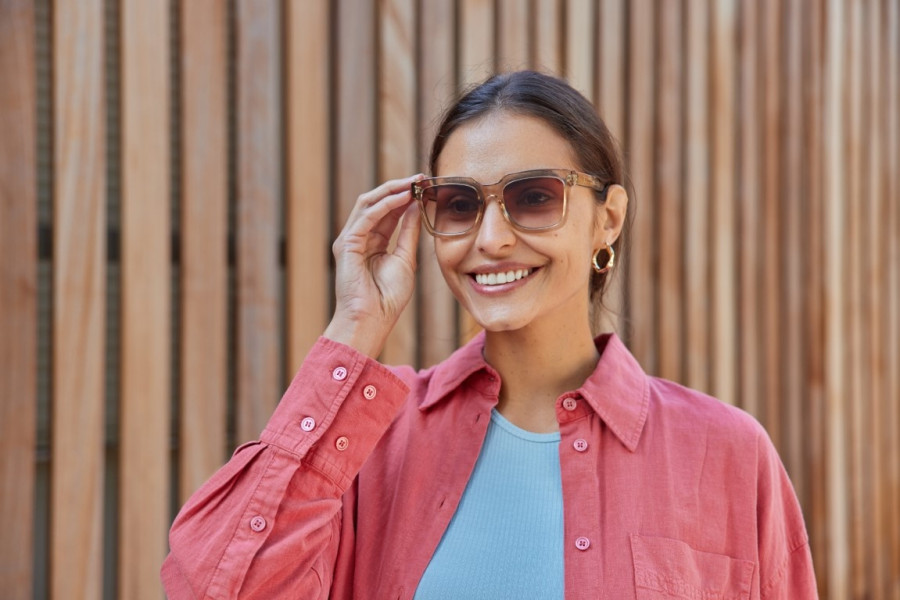 Satisfied fashionable brunette woman wears stylish sunglasses and pink shirt smiles toothily concent