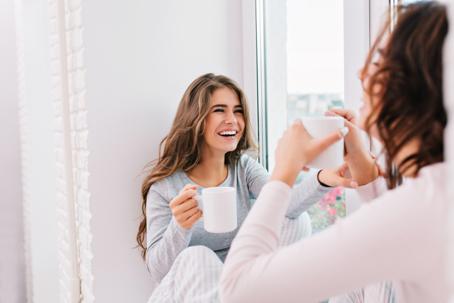 Portrait beautiful girl with long hair in pajama drinking tea on window in light room. She is smilin
