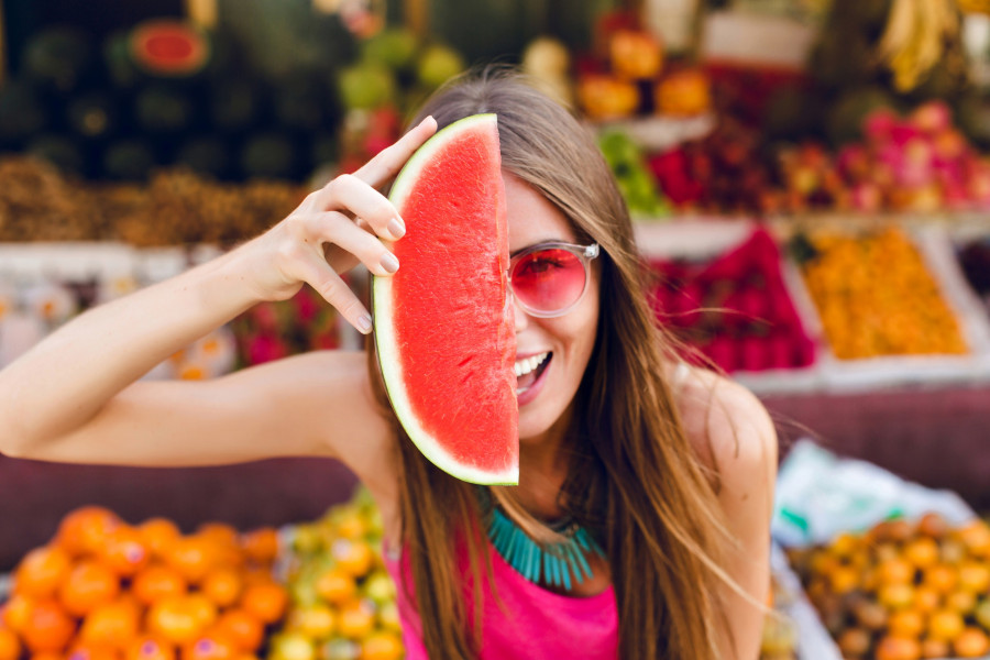 Closeup portrait of funny girl in pink sunglasses holding slice of watermelon on half face on tropical fruits background