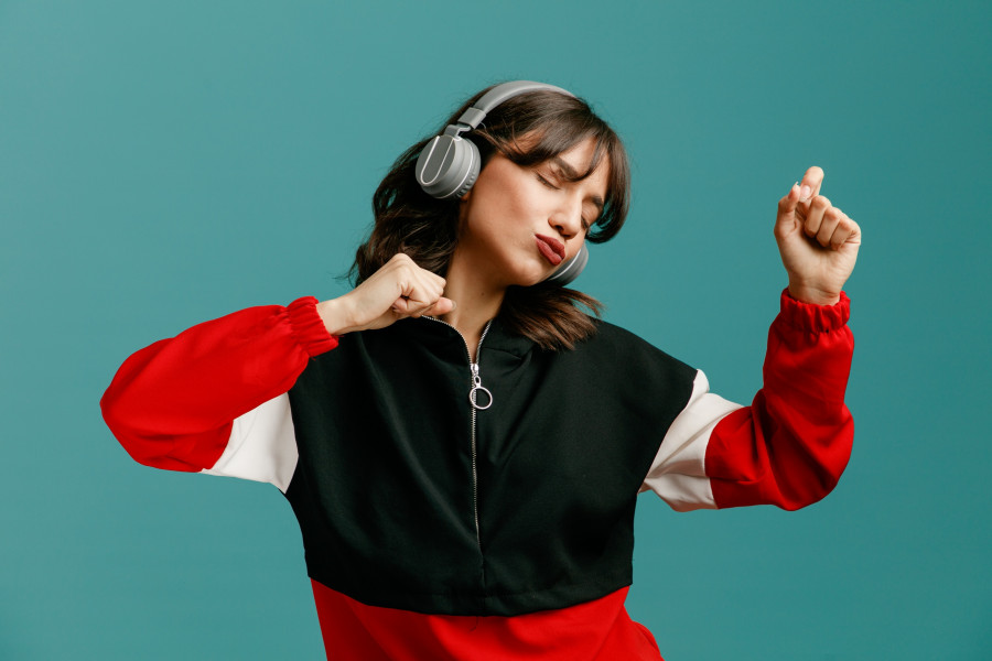 confident young caucasian woman wearing headphones keeping fists in air dancing with closed eyes isolated on blue background