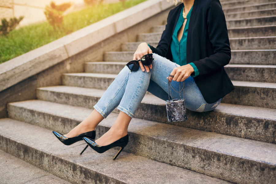young stylish beautiful woman sitting in street, wearing jeans, black jacket, green blouse, sunglasses, holding purse, elegant style, summer fashion trend, legs details, high heeled shoes, footwear