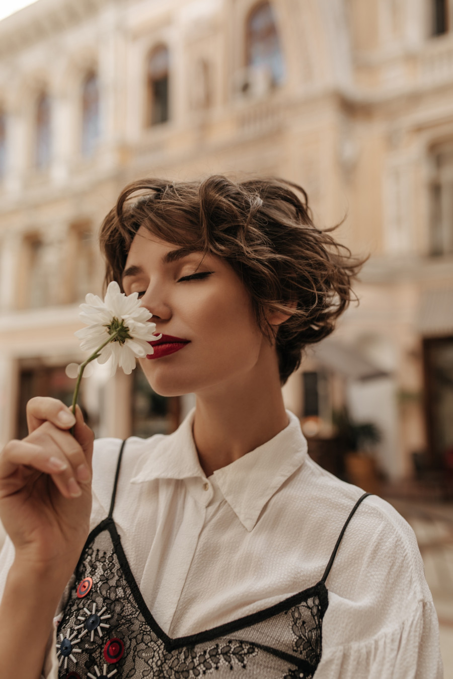 Wavy-haired girl in light shirt with black lace sniffing flower in city. Tender woman with red lips and short hair poses at street.