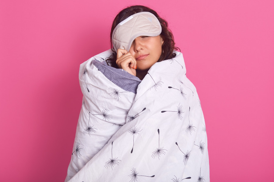 Close up portrait of brunette woman peeping from sleeping mask, does not want to wake up, keeps eyes closed, wearing white blanket, isolated over pink studio wall. People, lifestyle, morning concept.