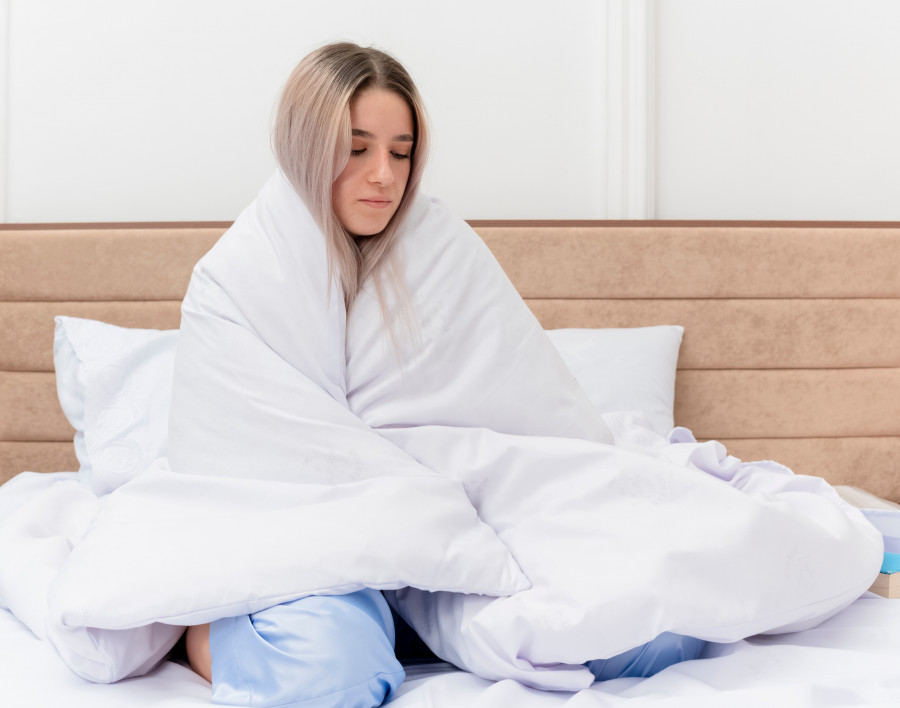 young beautiful woman in blue pajamas sitting on bed wrapping in blanket feeling unwell in bedroom interior on light background