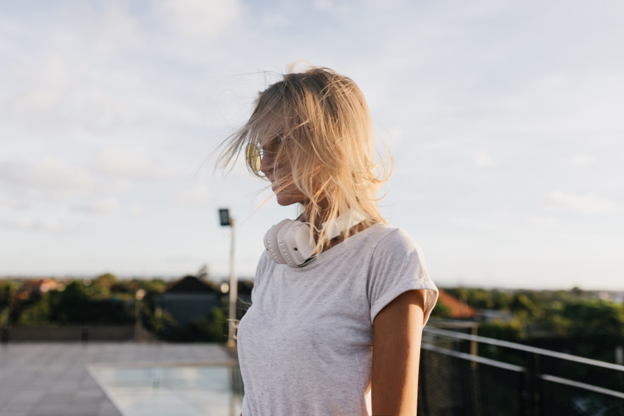 Pensive woman in white t-shirt looking away while walking around town in evening. Outdoor shot of stylish blonde girl in headphones posing on sky background.