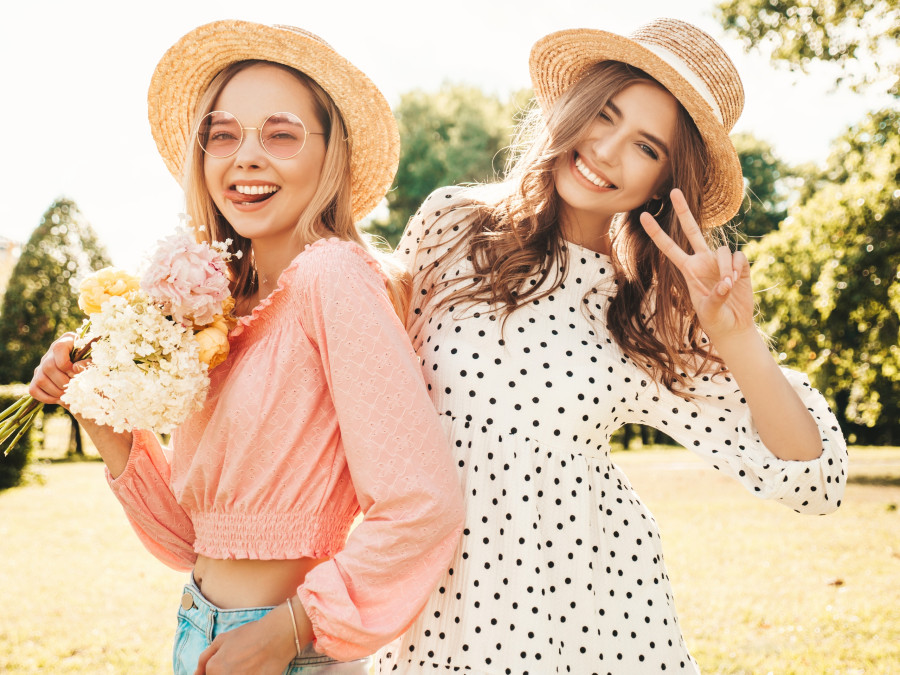 Two young beautiful smiling hipster woman in trendy summer sundress. Sexy carefree women posing in the park in hats. Positive models having fun and hugging at sunset. They holding flowers